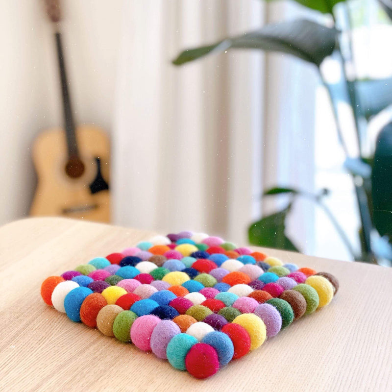 A handmade rainbow square felt ball pot trivet rests on a light wooden surface in a softly lit, homey environment, evoking a playful atmosphere. Crafted from 100% organic lamb wool, this fair-trade felt trivet features a colorful array of hand-stitched felt balls in shades of red, orange, yellow, green, blue, purple, and white.