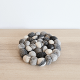 A round, thick wool felt pebble-stone pot trivet is displayed on a light-colored wooden surface against a plain white wall, exuding a calm and natural aesthetic. Handcrafted from 100% organic lamb wool, this unique felt trivet features closely arranged felt balls in varying shades of gray and white, resembling natural stones. This ethically made, organic piece showcases minimalist elegance and rustic charm, supporting Nepalese female artisans.