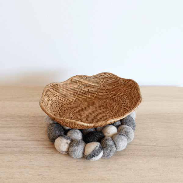 A handcrafted organic wool felt Pot Trivet rests on a light wooden surface against a plain white background, with an empty woven basket atop it. This thick, circular trivet, crafted from 100% organic lamb wool, features pebble-like felt balls in varying shades of gray and white. Ethically made by Nepalese female artisans, this unique piece adds organic texture and supports fair trade.