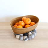 A thick, pebble-stone-like wool felt pot trivet rests on a light wooden surface against a plain white background, supporting a woven basket filled with vibrant oranges. This handcrafted trivet is composed of organic lamb wool felt balls in various shades of gray, resembling natural pebbles. Its ethically made design adds rustic charm and warmth, highlighting its fair-trade craftsmanship.