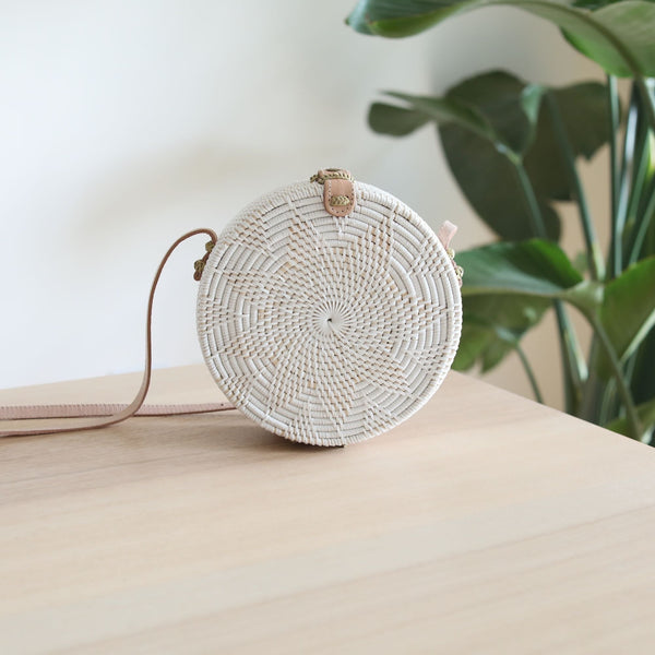A white Bali Star Round Rattan Crossbody Bag rests on a wooden surface, surrounded by a plant in the background. The bag features a round shape with a natural star design pattern, an adjustable leather strap, and is made from high-quality rattan materials.