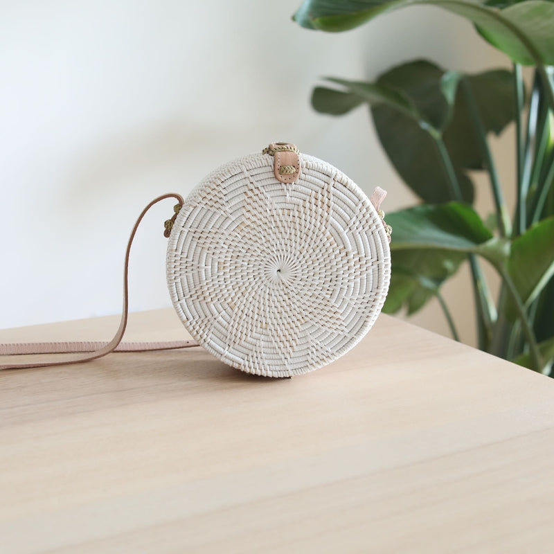 A white Bali Star Round Rattan Crossbody Bag rests on a wooden surface, surrounded by a plant in the background. The bag features a round shape with a natural star design pattern, an adjustable leather strap, and is made from high-quality rattan materials.