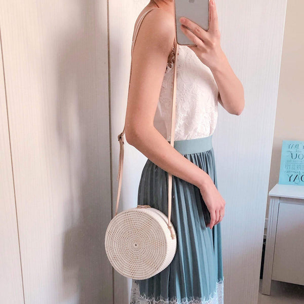 A woman poses for the camera, wearing a white top and light blue skirt, holding her phone in one hand. She wears a round woven rattan crossbody bag on her left shoulder with an adjustable leather strap. The bag features a unique star design and appears to be made of natural rattan materials. It is white in color, round in shape, medium in size, and has a textured appearance.
