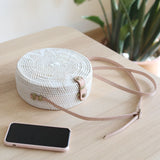 A round, white Bali Star Round Rattan Bag is showcased on a wooden surface next to an iPhone, featuring a woven rattan texture with a prominent star design. The bag has an adjustable leather strap and closure mechanism, accompanied by a plant in the background.